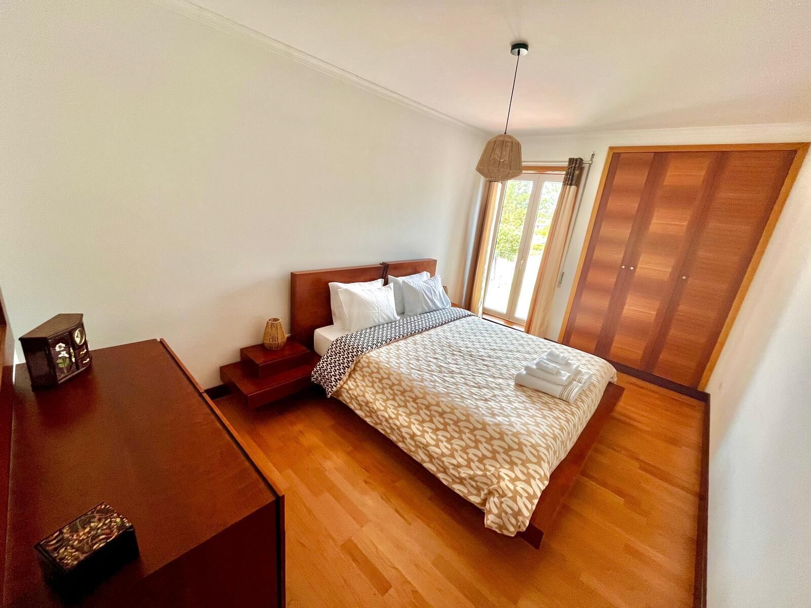 A bedroom with a wooden bed, a bedside table, a lamp, and a view of the outdoors through a sliding door.