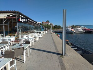 A waterfront café with outdoor seating, a clear view of the marina, and a sign reading "Aqua By the sea". - Apartment 3 Rooms, Nearby Casino, Cliff, Port, Aquarium, Beach, Ovidius Square (Constanta)