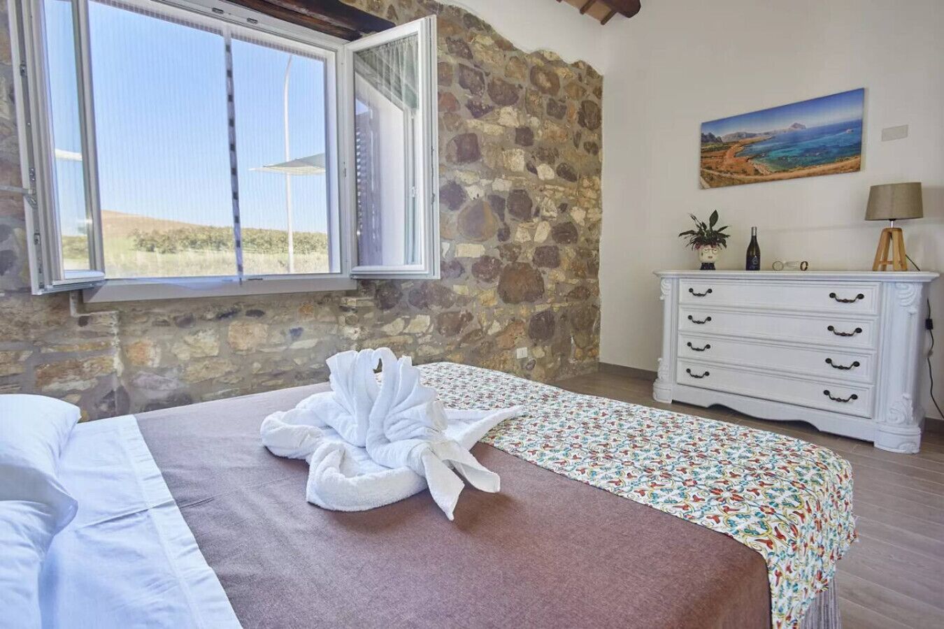 A bedroom with a stone wall, a bed with a white towel, a dresser, and a painting of a landscape.