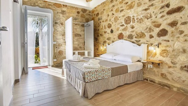 A bedroom with a stone wall, a bed with white bedding, a bedside table with a lamp, and a view through a glass door.