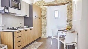 A compact kitchen with a microwave, stove, and wooden cabinets. A dining area with a table and chairs is visible through a stone archway.