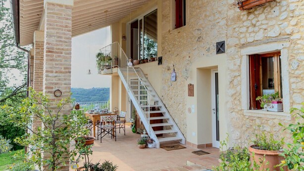 A stone building with a wooden staircase leading to a balcony. There is a table and chairs outside, and potted plants are visible.