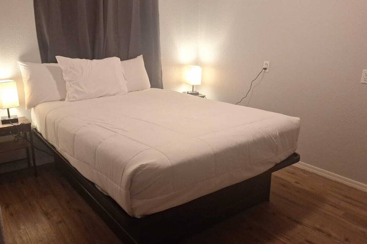 A neatly made bed with a white comforter and pillows, two bedside lamps, a wooden nightstand, and a power outlet on the wall.