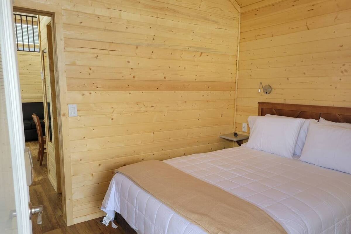 A wooden room with a bed, a nightstand, and a door leading to another room.
