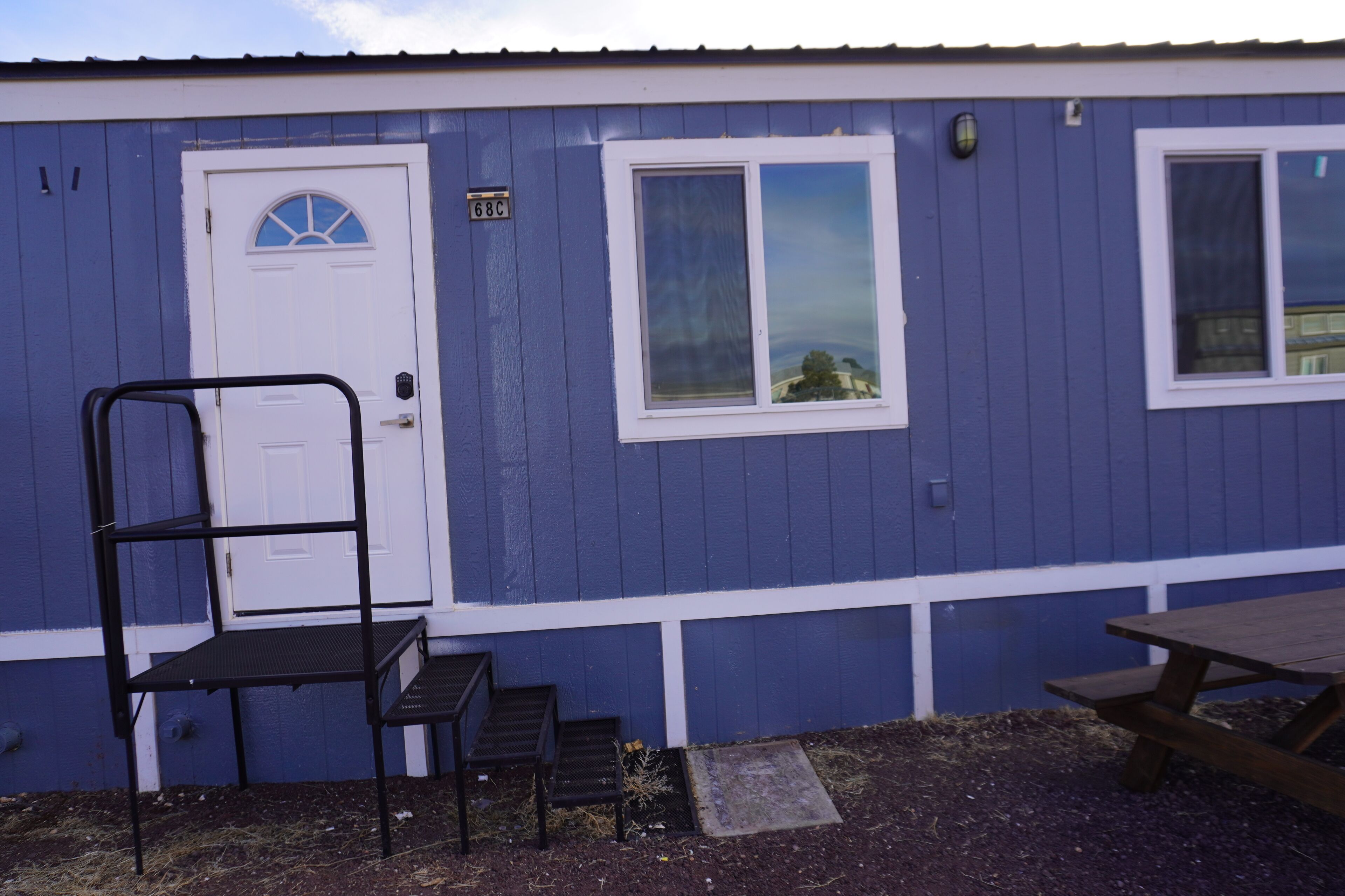 A small, single-story building with a white door and a window. There is a black metal bench and a picnic table outside.