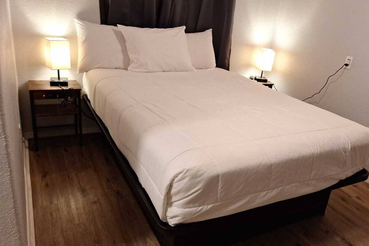 A neatly made bed with a white duvet, two bedside lamps, and a power outlet on the wall.