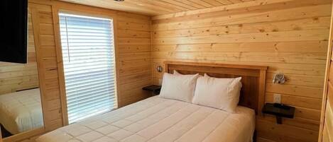 A wooden cabin bedroom with a large bed, a nightstand, a TV mounted on the wall, and a window with blinds.