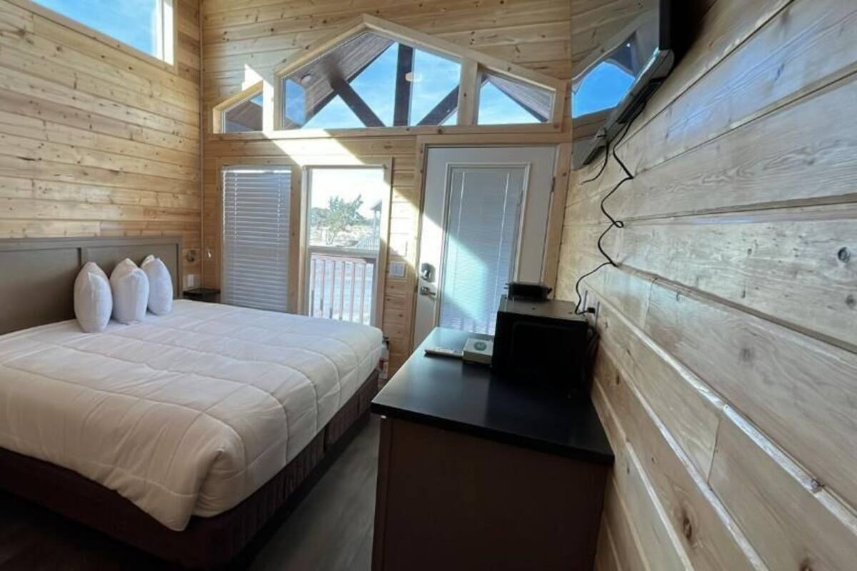 A wooden cabin bedroom with a bed, a desk, and a window overlooking the outside.
