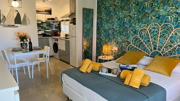 A modern hotel room with a bed, a dining area, and a kitchenette.