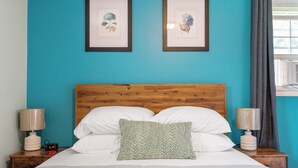 A neatly made bed with white linens and a green pillow, flanked by two bedside tables with lamps, against a teal wall with two framed botanical prints.