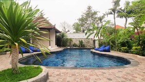 Una piscina con borde de piedra, rodeada de plantas tropicales y un edificio con techo de tejas al fondo.