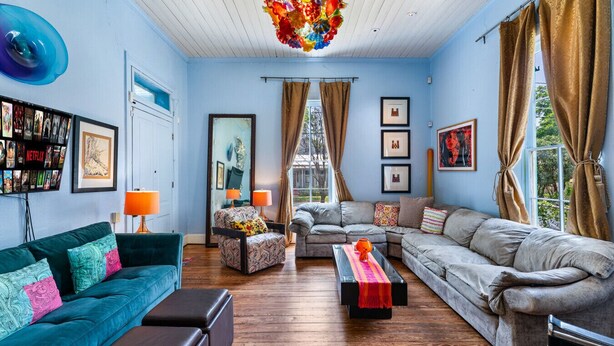 Ein Wohnzimmer mit einem türkisfarbenen Sofa, einem grauen Sofa und einem gemusterten Sessel. Zwei Lampen, eine farbenfrohe Kronleuchter und ein Couchtisch mit roter Decke sind ebenfalls vorhanden.