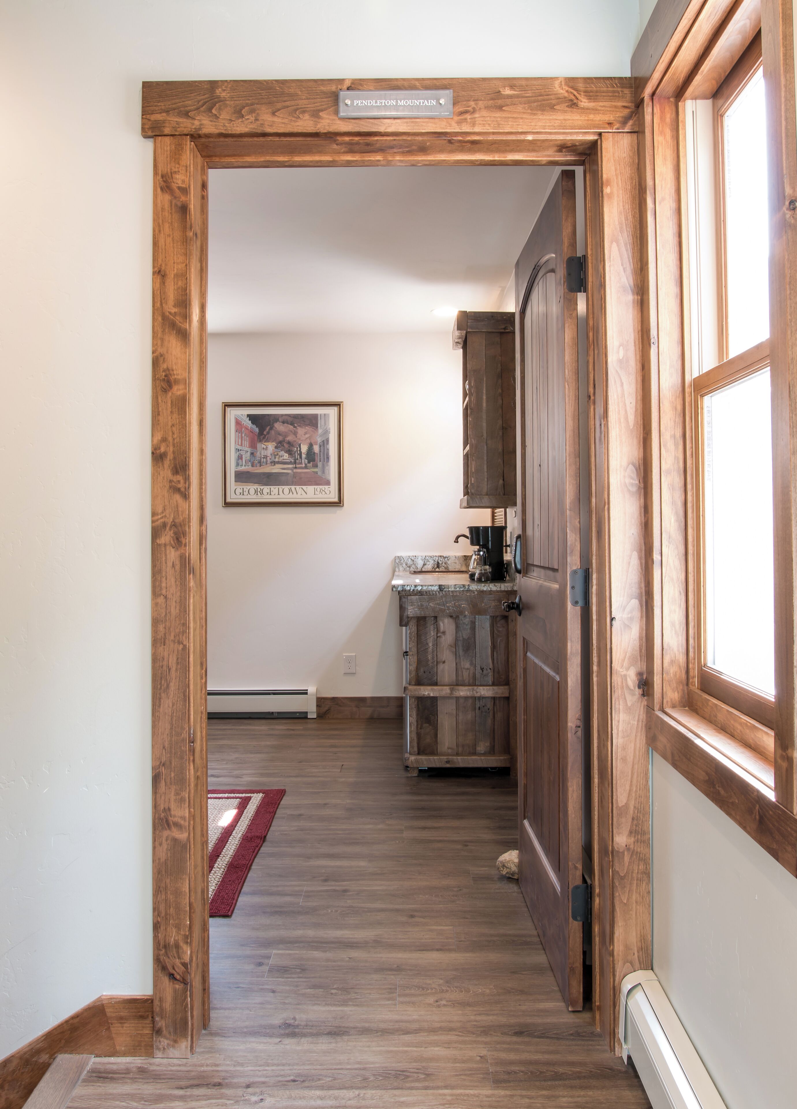 Un passage encadré de bois mène à une cuisine au décor rustique, avec un tableau encadré accroché au mur et un tapis rouge sur le sol.