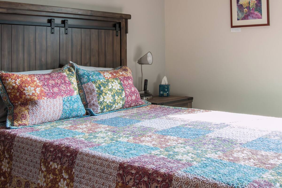 A bedroom with a wooden headboard, a colorful quilt, a bedside lamp, and a framed picture on the wall.