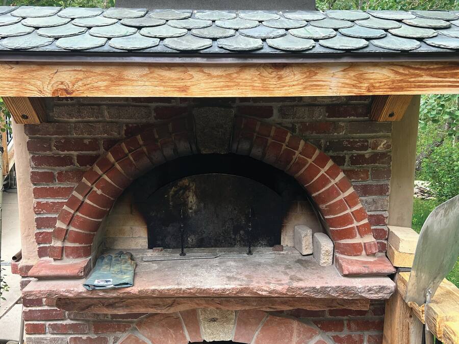 Een buitenoven voor pizza, gebouwd van baksteen, met een schuin dak, een vuurruimte en een houtopslag.