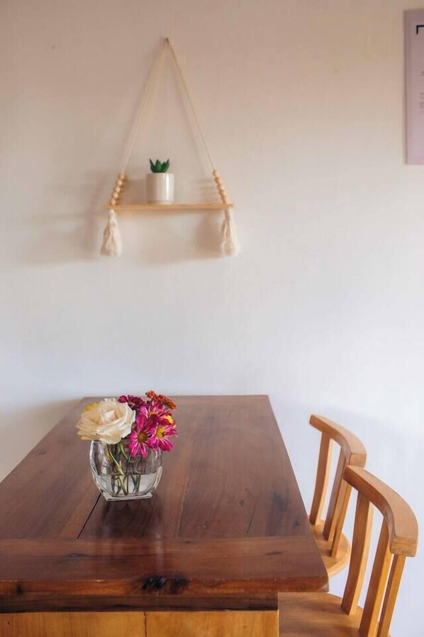 Una mesa de comedor de madera con un florero de vidrio, una estantería de macramé con una planta pequeña y sillas de madera.