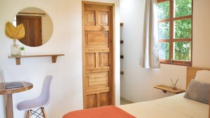 A bedroom with a wooden door, a round mirror, a bed with a white and orange bedspread, a bedside table with a lamp, and a window with curtains.