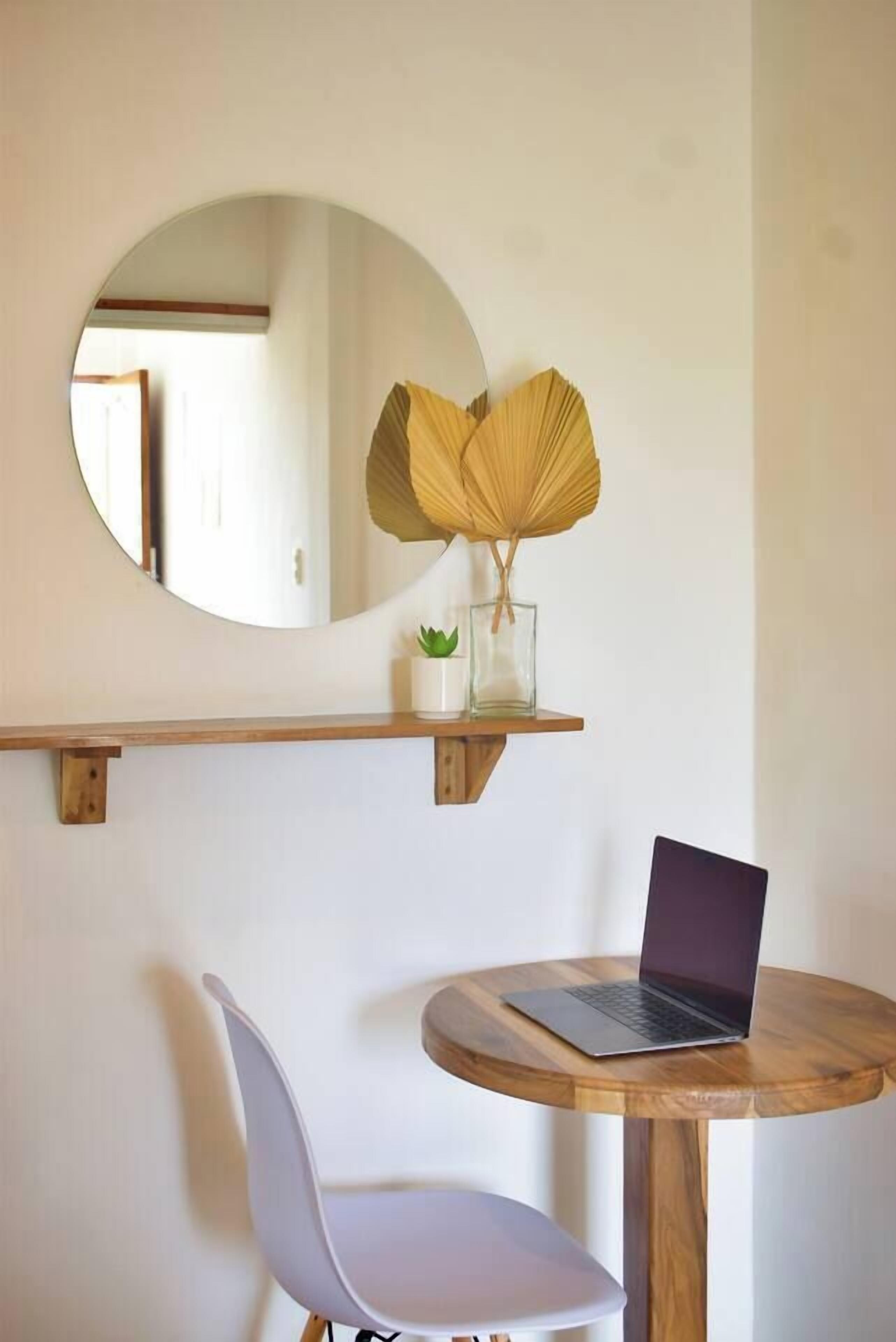 Een ronde houten tafel met een laptop, een witte stoel, een spiegel en een plankje met een plant en een decoratief object.