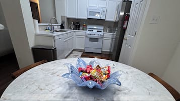 A kitchen with white cabinets, a microwave, and a stove. There is a round marble table with a clear bowl containing snacks.