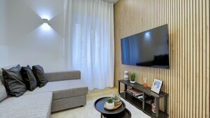 A modern living room with a grey sofa, black cushions, a small black table with a tray, a flat-screen TV mounted on a wooden wall, and a ceiling light. - Elegance and comfort in San Giovanni, 3 minutes from metro and ideal for vacation. (Roma)