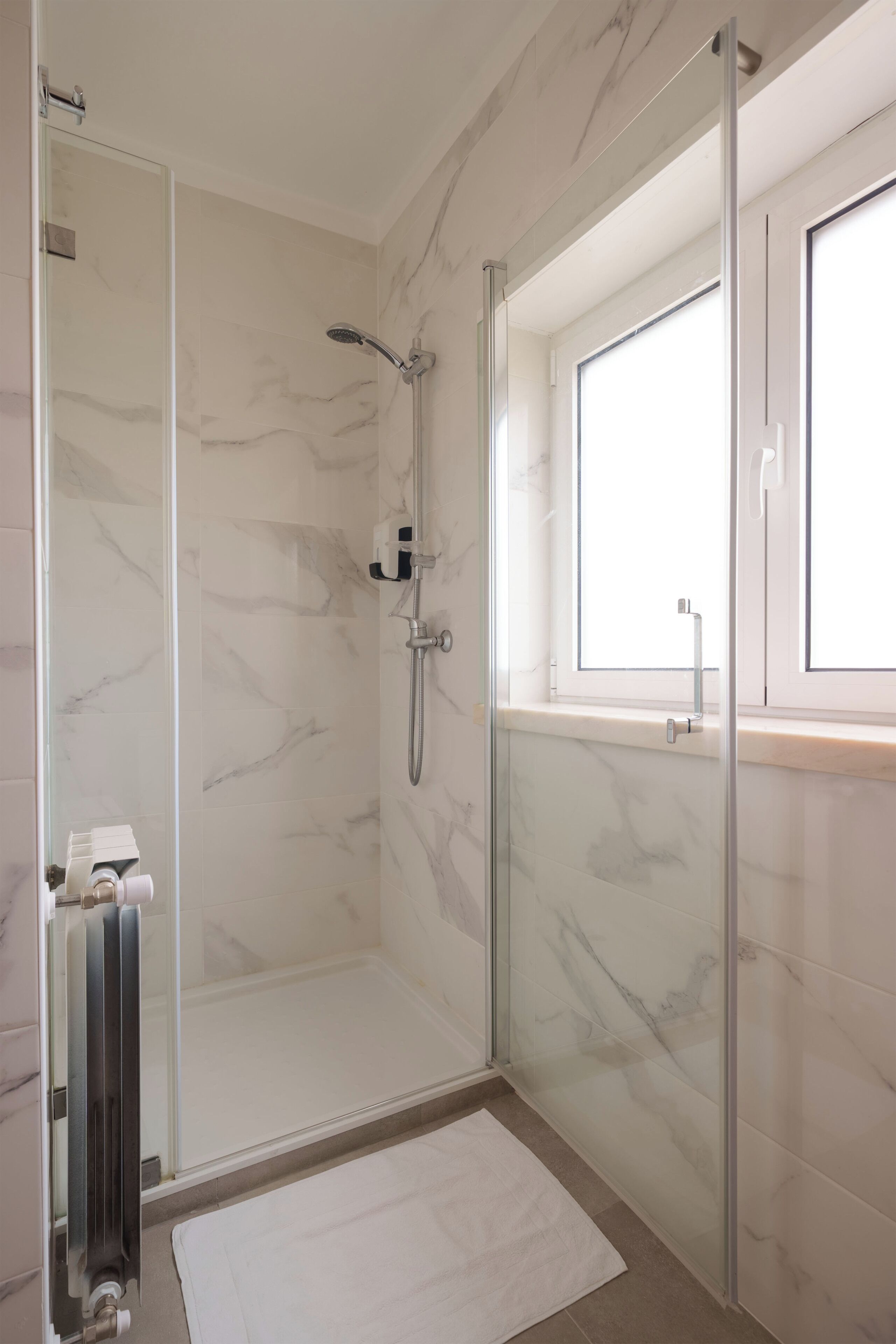A modern bathroom with a glass-enclosed shower, marble tiles, and a window.