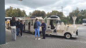 Un train d’époque, avec des passagers montant à bord, est garé sur une zone pavée, avec des arbres en arrière-plan.