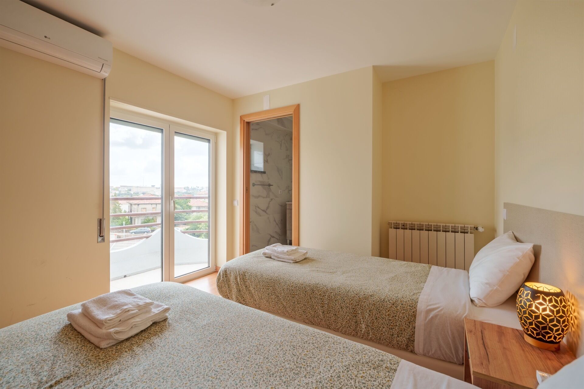 A hotel room with two beds, a radiator, a wooden nightstand with a lamp, and a sliding glass door leading to a balcony.
