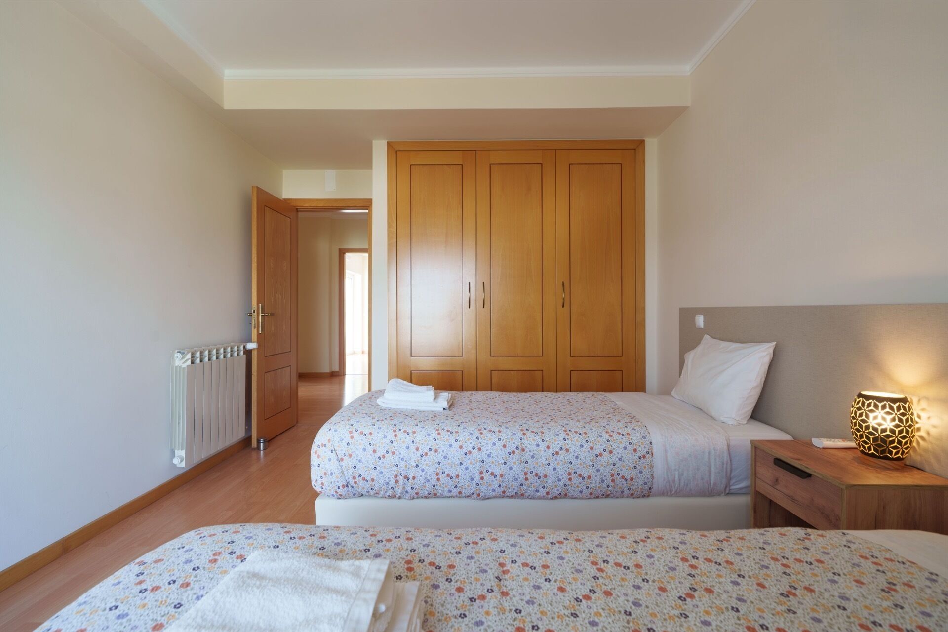 A bedroom with two beds, a wardrobe, a bedside table with a lamp, and a radiator.