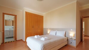 A bedroom with a large bed, wooden wardrobe, bedside lamps, and a bathroom visible through an open door.