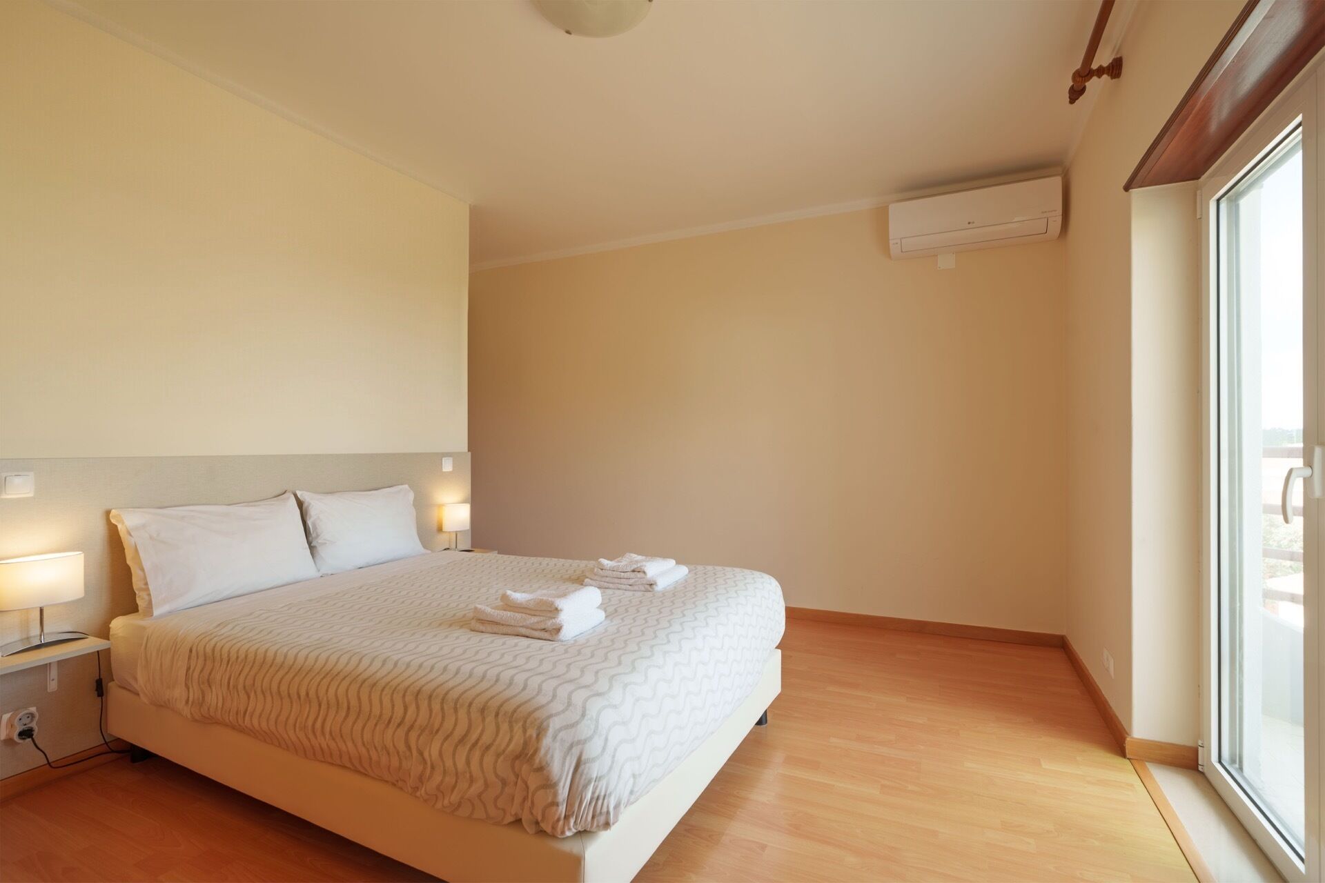 A hotel room with a large bed, bedside lamps, an air conditioning unit, and a window with a view.