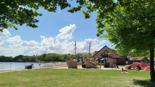 A park with a wooden play structure, a small building, and a lake. - Vacation semi-detached house Tannenhuuske at the bathing lake Aurich-Tannenhausen East Frisia (Aurich)