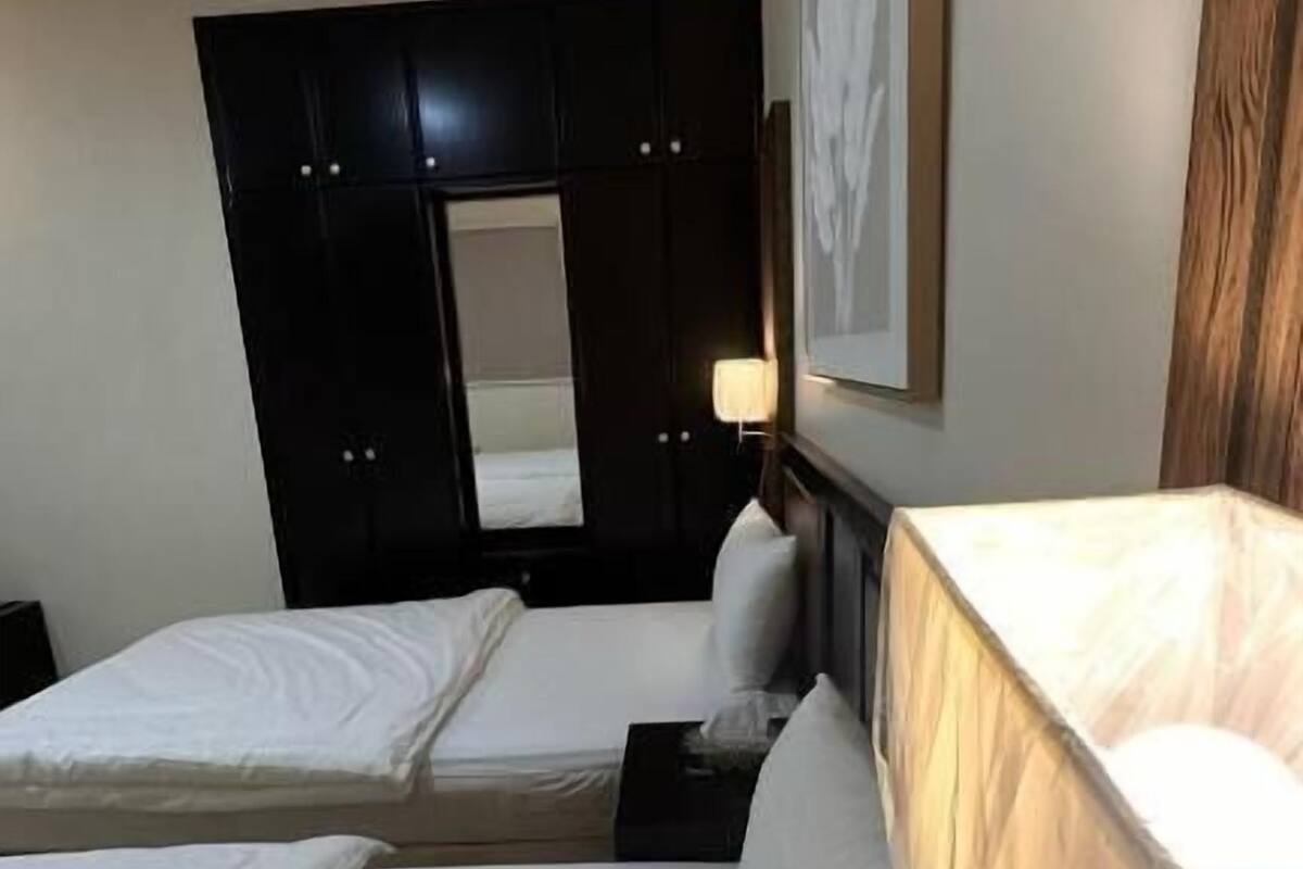 A hotel room with two single beds, a dark wooden wardrobe, a bedside table with a lamp, and a framed picture on the wall.