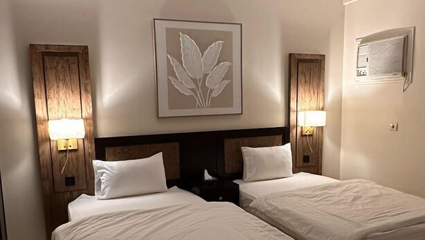 A hotel room with two beds, a wooden headboard, wall-mounted lamps, and a framed artwork. - AlShowaish Apartments (Taif)