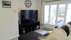 A living room with a large flat-screen TV, a clock, a potted plant, and a view of the street through a large window.