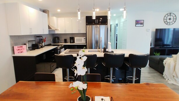 A modern kitchen with white cabinets, a wooden dining table, and black chairs. There is a sign on the wall that reads "HOME SWEET HOME."