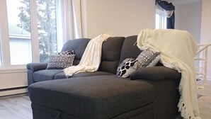 A dark grey sectional sofa with a white throw blanket and patterned pillows, positioned in a room with wooden flooring and a large window.