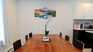 A dining area with a wooden table, six chairs, a vase with flowers, and a wall-mounted painting of a mountain landscape.
