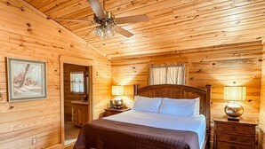 Een slaapkamer in een houten hutje, met een bed, twee nachtkastjes, een plafondventilator en een schilderij aan de muur.