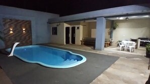 Una piscina de borde ondulado, rodeada por un patio de concreto. Dos puertas conducen a un espacio cubierto con una mesa y sillas.