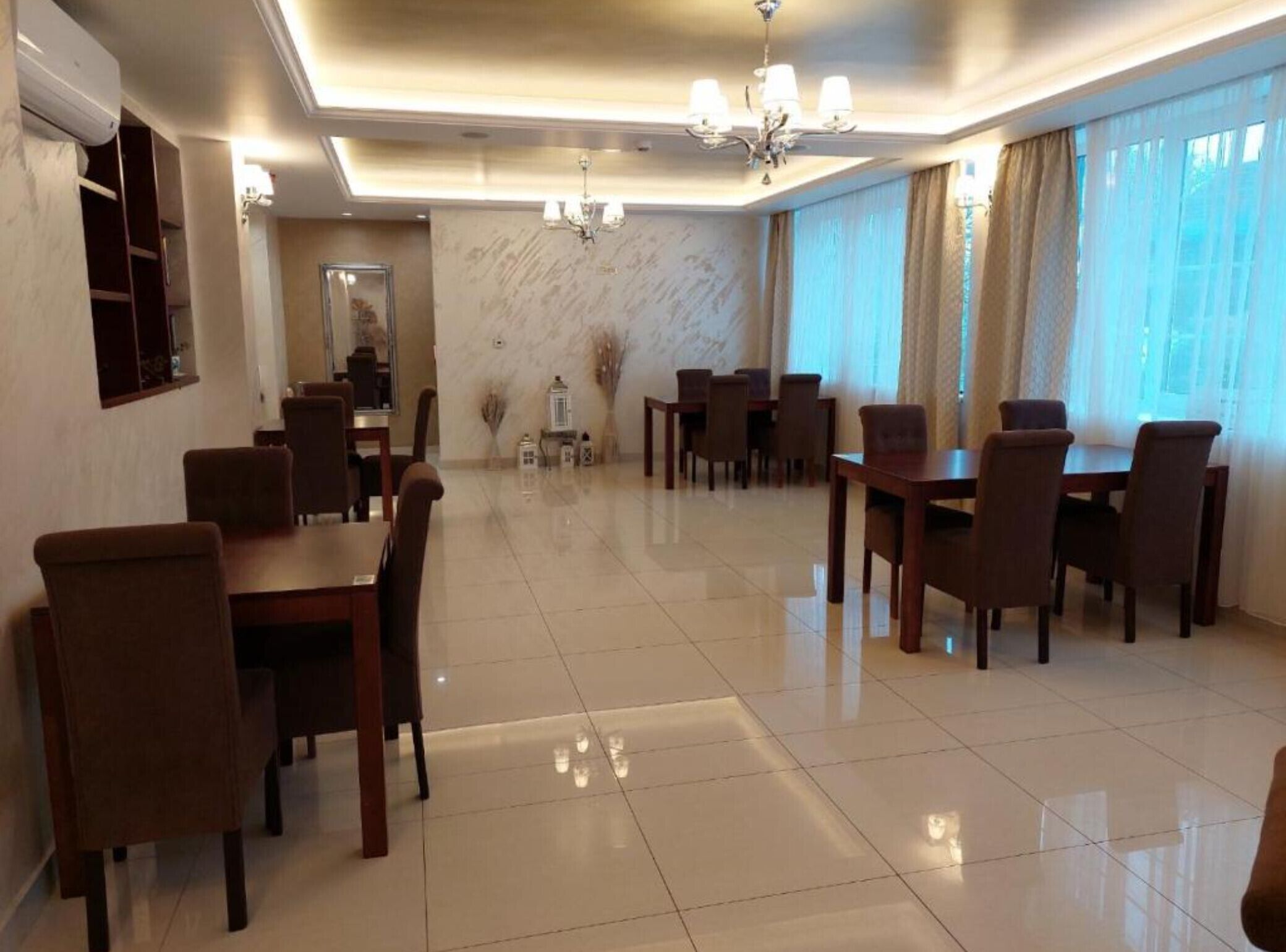 A spacious dining area with multiple tables and chairs, a chandelier, and large windows with curtains.