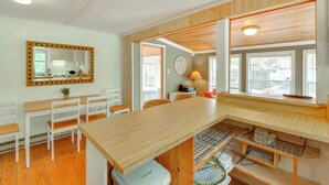Una cocina con encimera de madera, sillas blancas y una mesa de comedor. Hay un espejo en la pared y una ventana con persianas.