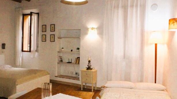 A bedroom with two beds, a nightstand, a lamp, a shelf with books and a plant, and a window with curtains. - "Lovely Home" Cozy Bike Friendly Holiday Home with Wi Fi (Buonconvento)