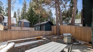 A backyard with a stone barbecue area, a wooden table, and a wooden fence. - Brooklyn Cabins (Big Bear Lake)