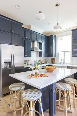 Une cuisine moderne avec des meubles de couleur bleu foncé, un réfrigérateur en acier inoxydable, un grand îlot central doté de tabourets et un éclairage suspendu.