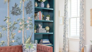Ein Wohnzimmer mit einem türkisblauen Bücherregal, einem rosa Sofa, einem runden Holztisch und einem großen Fenster mit floralen Vorhängen.
