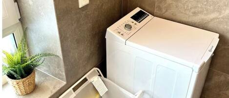 Une petite buanderie avec un lave-linge blanc, un panier à linge en plastique et une plante en pot posée sur une étagère.