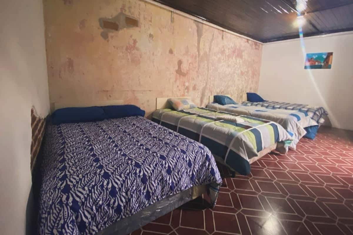 A room with three beds, each with patterned bedding. The walls are worn, and the floor has a geometric tile pattern.