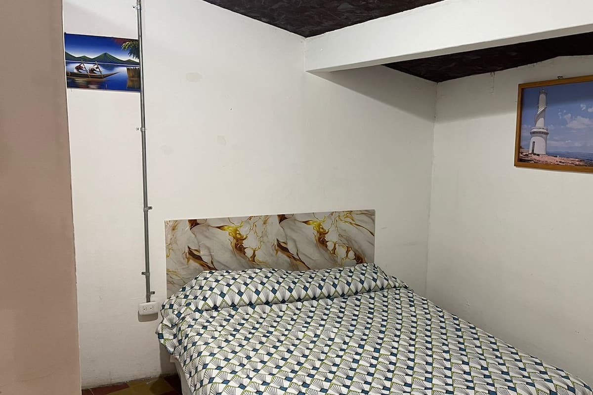 A small, sparsely furnished room with a bed, a single wall-mounted picture, and a visible electrical outlet.