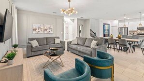 Een moderne woonkamer met grijze banken, een salontafel en teal fauteuils. Er is een eethoek met een tafel en stoelen, en een keuken op de achtergrond.