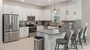Een moderne keuken met witte kasten, roestvrijstalen apparatuur, een centraal kookeiland met barstoelen en hanglampen.